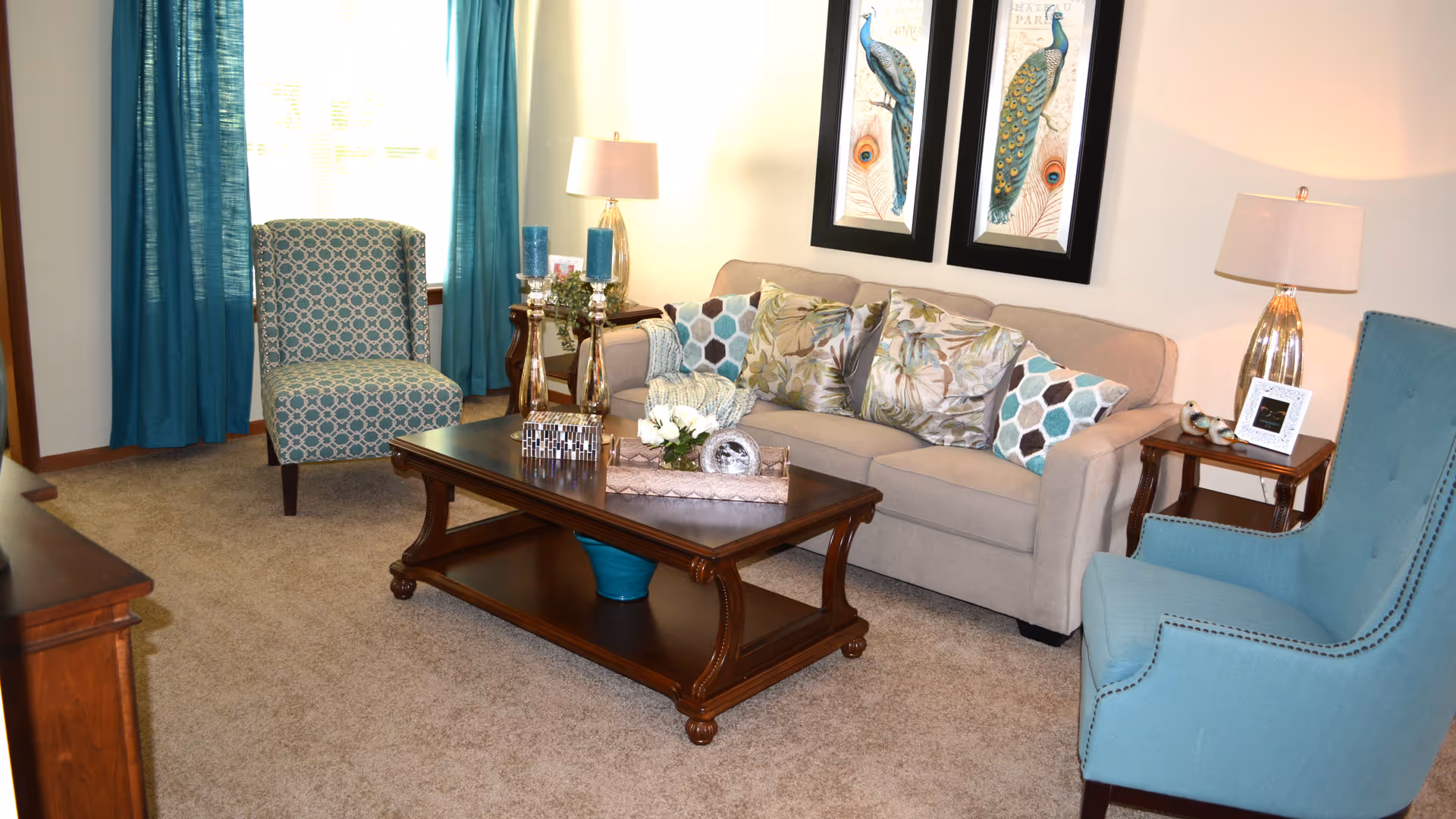A cozy living room with a beige sofa adorned with patterned cushions, two blue armchairs, a wooden coffee table with decorative items, two framed peacock prints on the wall, and two side tables each with a lamp. Blue curtains frame a window in the background.