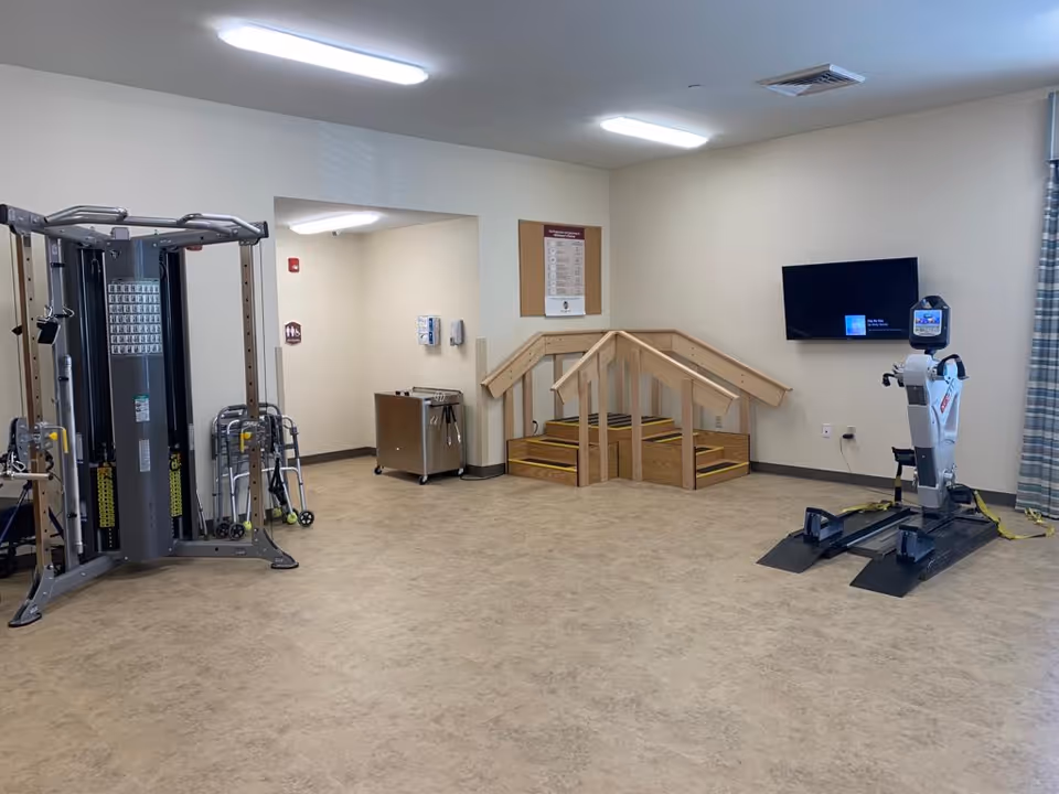 A rehabilitation or physical therapy room with exercise equipment including a multi-gym machine on the left and a seated exercise machine on the right. In the center back, there is a small set of wooden stairs with handrails for physical therapy exercises. A flat-screen TV is mounted on the wall to the right, and there is a cart and a bulletin board on the back wall. The room has beige walls and a light-colored floor.
