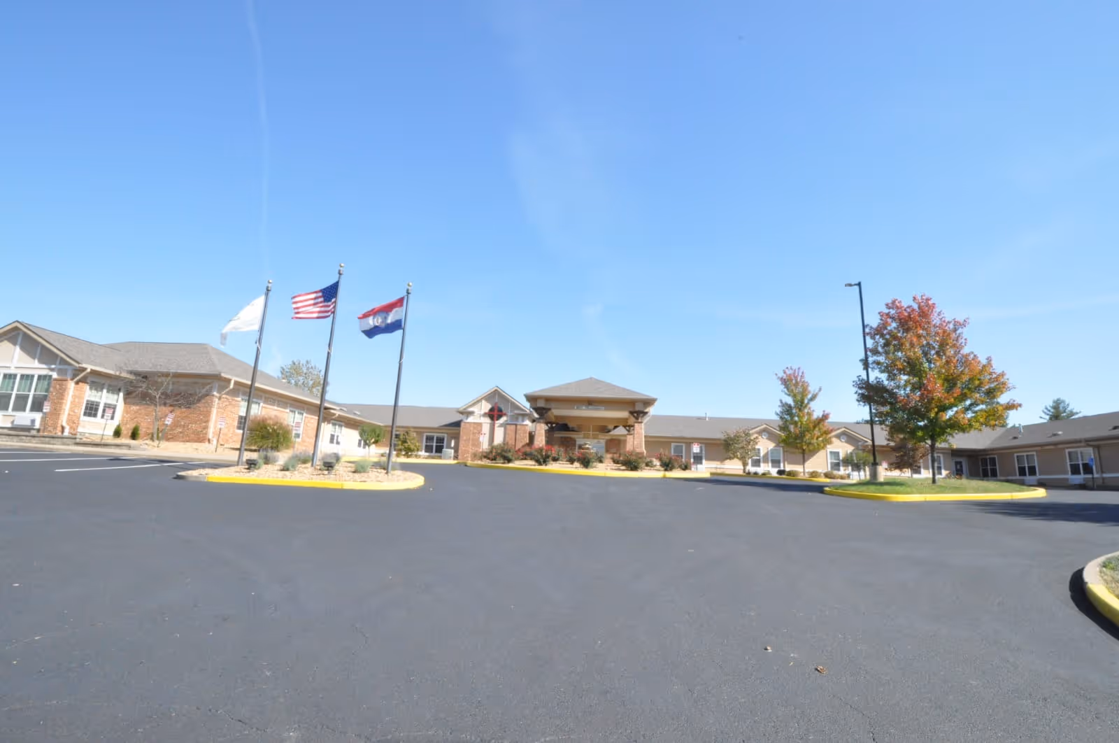 Wide front view of a single-story brick nursing facility with flagpoles and a circular driveway under a clear blue sky.