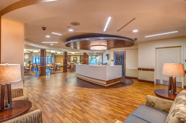 A spacious and well-lit interior area of a senior living facility featuring a central reception desk with a circular ceiling design above it. The room has wooden flooring, modern lighting fixtures, and comfortable seating areas with lamps on side tables. In the background, there are dining tables and chairs arranged neatly, indicating a communal dining space.