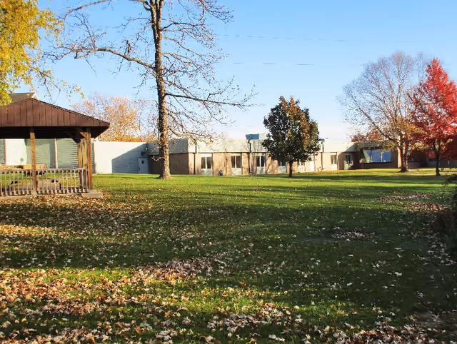 A grassy outdoor area with scattered fallen leaves, a wooden gazebo on the left, several trees with autumn foliage, and a single-story building in the background under a clear blue sky.