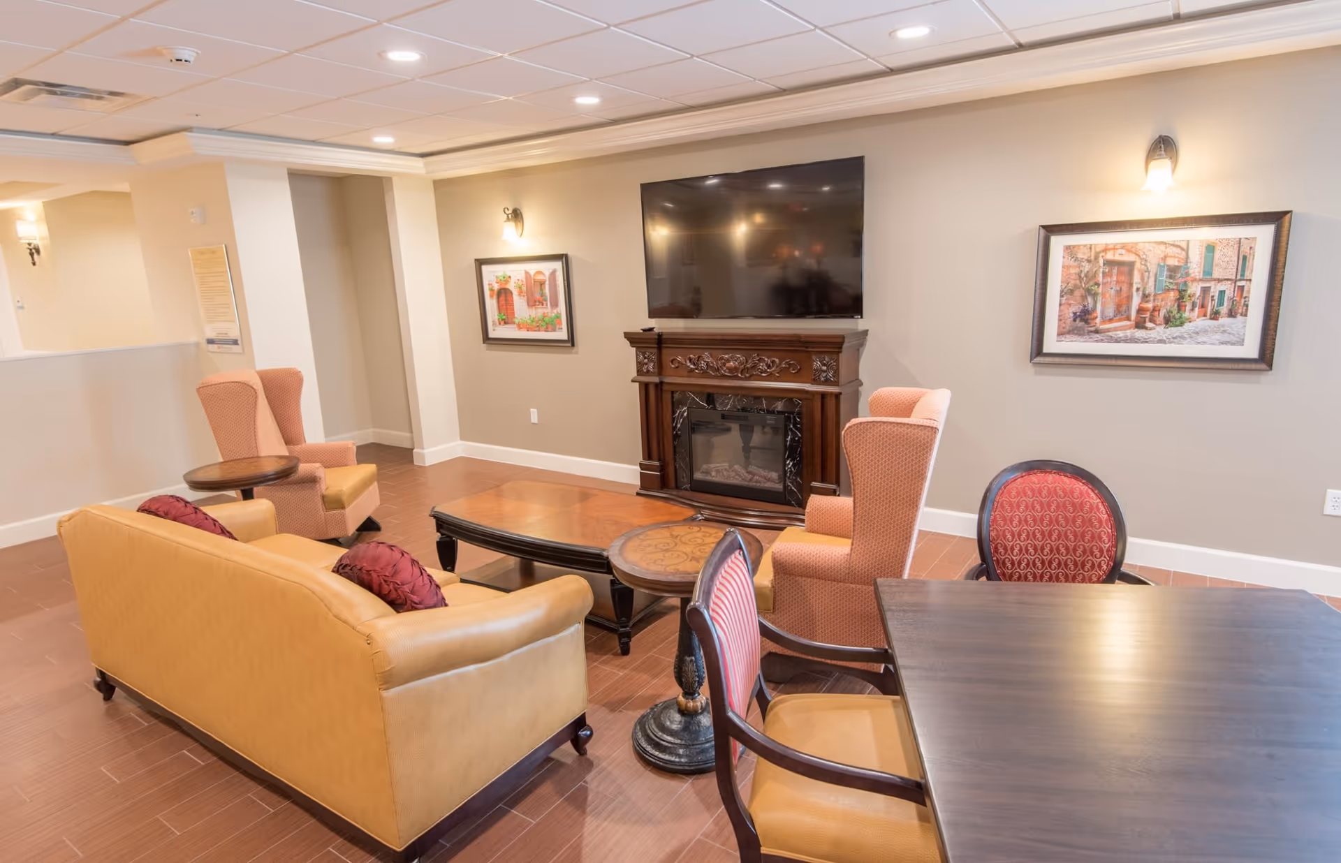 A cozy living room area with a tan leather sofa, two upholstered armchairs, a wooden coffee table, and a small round side table. A flat-screen TV is mounted above a decorative wooden fireplace. The walls are adorned with framed artwork and wall sconces, and the floor is covered with wood-like tiles.