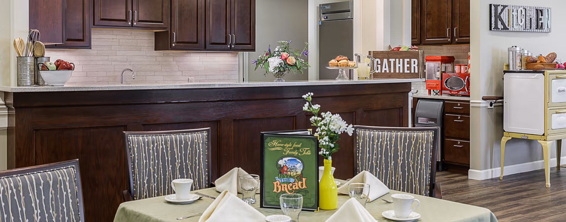 A dining area with a table set for four, featuring white cups, glasses, and folded napkins. Behind the table is a kitchen counter with dark wood cabinets, a stainless steel refrigerator, and various kitchen items including a 'GATHER' sign and a vase with flowers. On the right side, there is a vintage-style stove and a wall decoration spelling 'KITCHEN'.