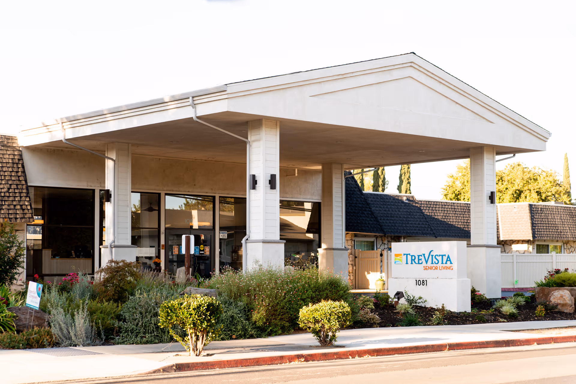 Exterior view of the entrance to TreVista Senior Living facility with a covered driveway, landscaping with bushes and flowers, and a sign displaying the facility name and address 1081.