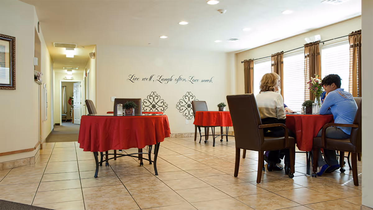 A bright dining area with tiled floors and cream-colored walls. There are round tables covered with red tablecloths and brown chairs around them. Two people are seated at one of the tables near large windows with brown curtains. The wall has decorative metal art and a quote that reads 'Live well. Laugh often. Love much.'