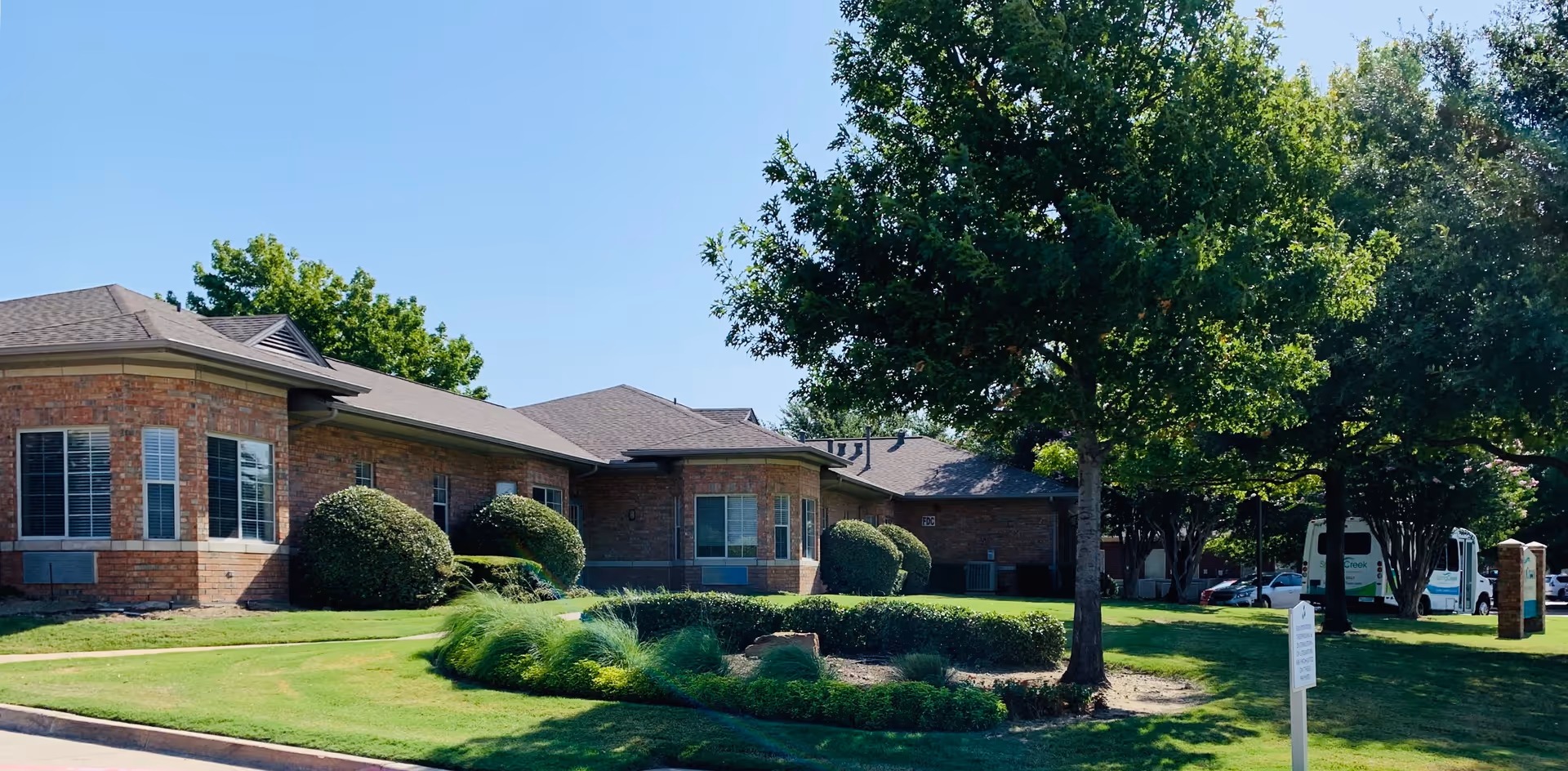 Brick single-story assisted living building with manicured lawn, shrubs, trees, and a parked shuttle bus at the driveway.