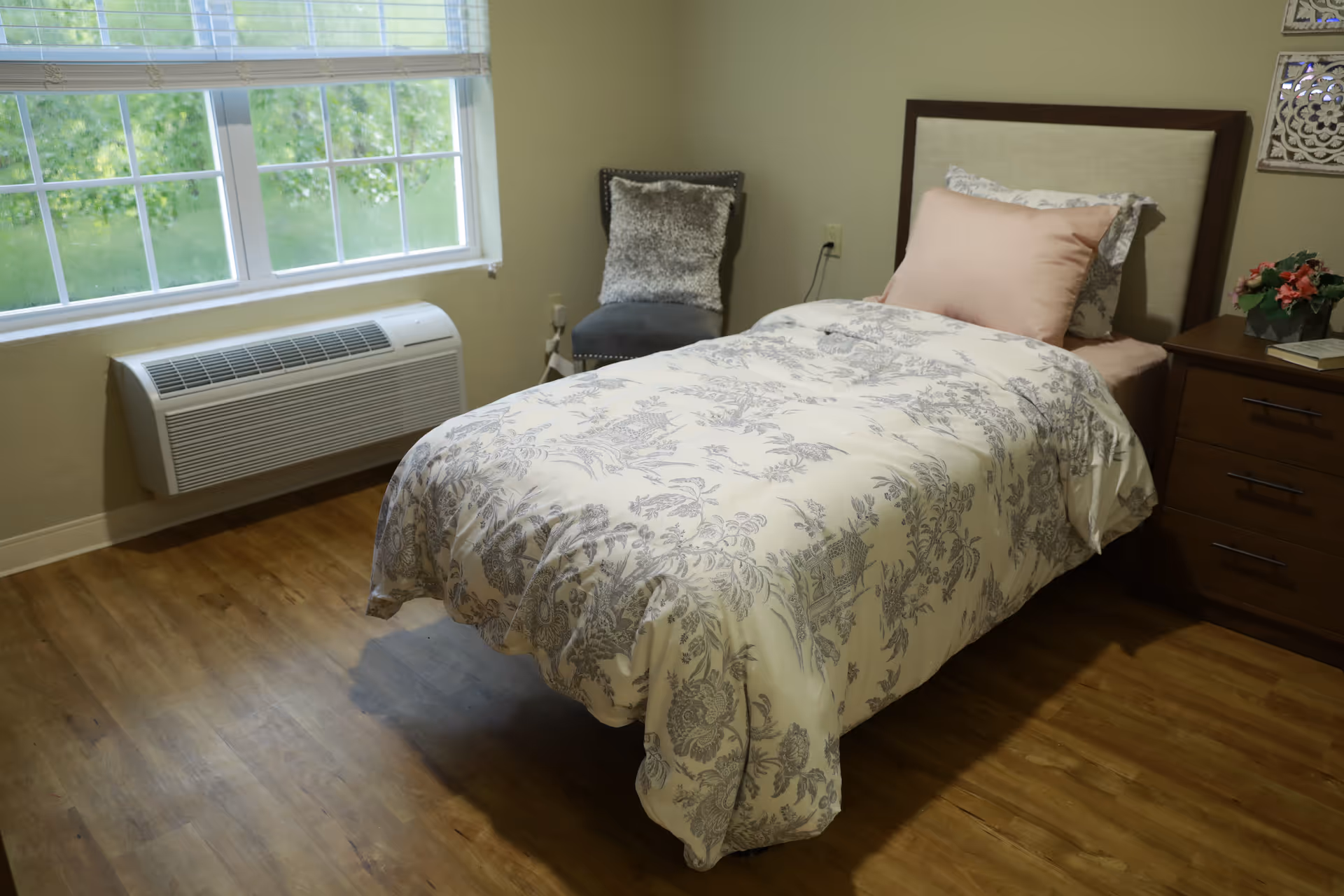 A cozy bedroom with a single bed covered in a white and gray floral patterned comforter and a pink pillow. Next to the bed is a wooden nightstand with a small plant and a book. A gray chair with a fluffy pillow is placed near a large window with white blinds, and a wall-mounted air conditioning unit is below the window. The room has wooden flooring and light-colored walls.