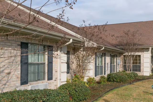 Exterior view of a single-story brick building with multiple windows featuring black shutters. The building is surrounded by trimmed bushes and small trees with no leaves, under a partly cloudy sky.
