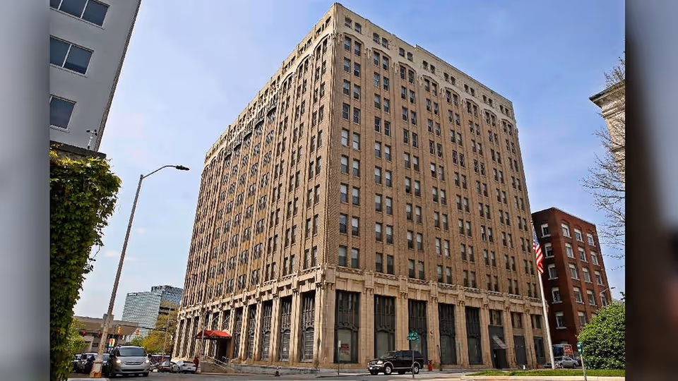 Exterior view of a tall, multi-story brick building on a city street corner under a clear blue sky. Several cars are parked and driving nearby, and other buildings are visible in the background.