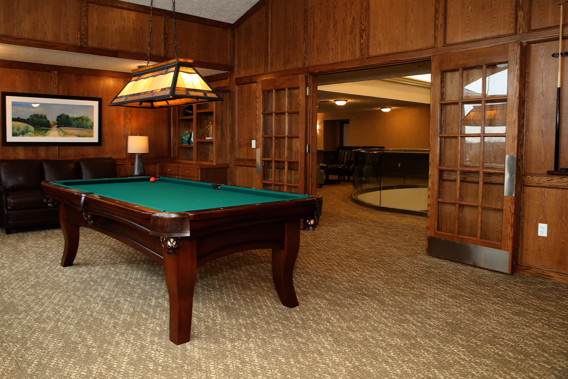 A room with a green felt pool table in the center, surrounded by wood-paneled walls. There is a leather couch against the wall with a framed landscape painting above it, and a lamp on a side table. The room has a carpeted floor and a hanging light fixture above the pool table. Double wooden doors with glass panes lead to another carpeted area with seating and a circular glass railing.