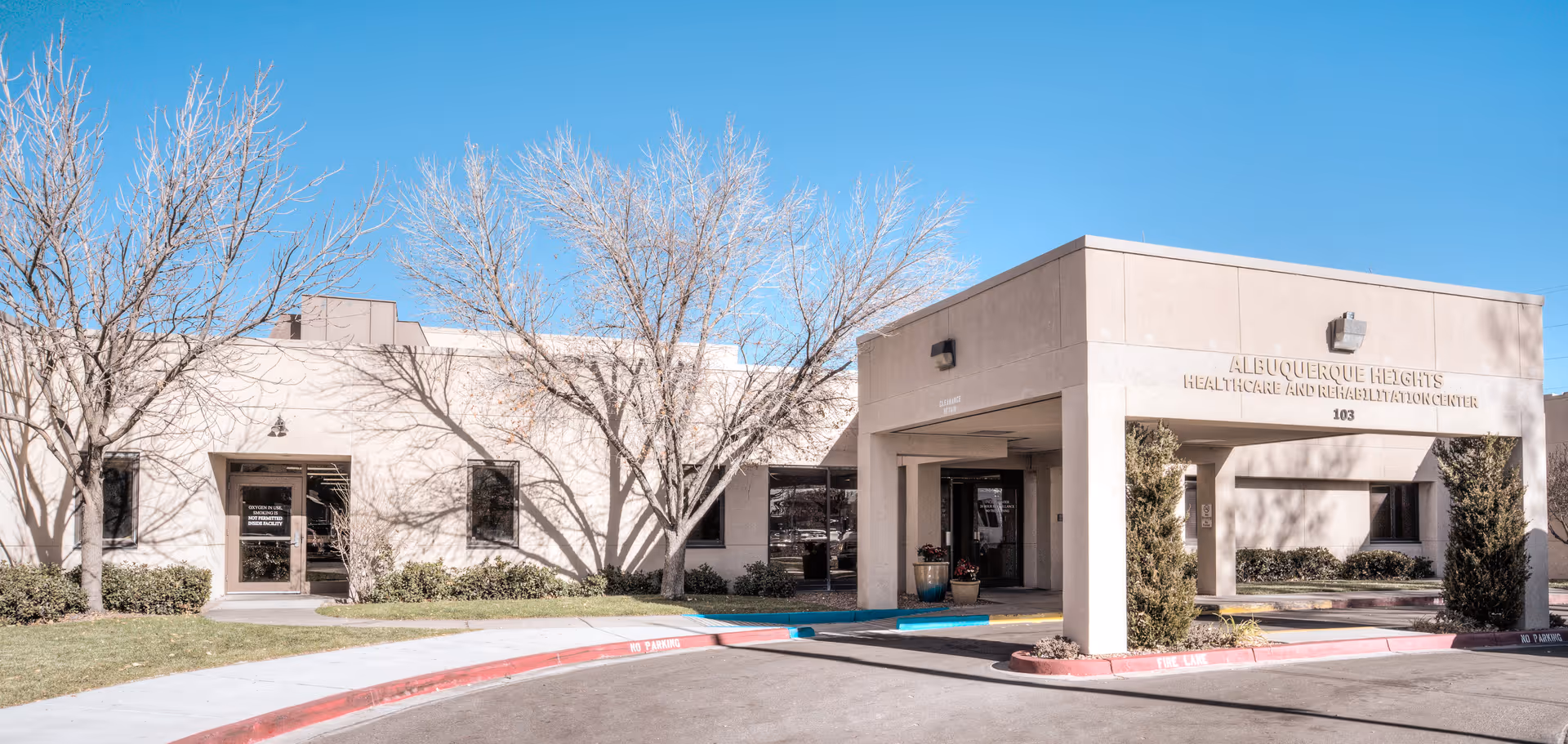 Exterior view of Albuquerque Heights Healthcare and Rehabilitation Center building with a covered entrance, leafless trees, and a clear blue sky.
