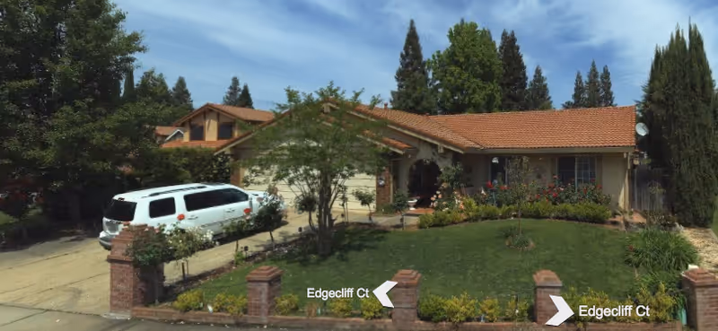 Front exterior view of a single-story house with a red-tiled roof, a green lawn, and a driveway with a white SUV parked. The house is surrounded by trees and bushes, and there is a brick fence with pillars along the front.
