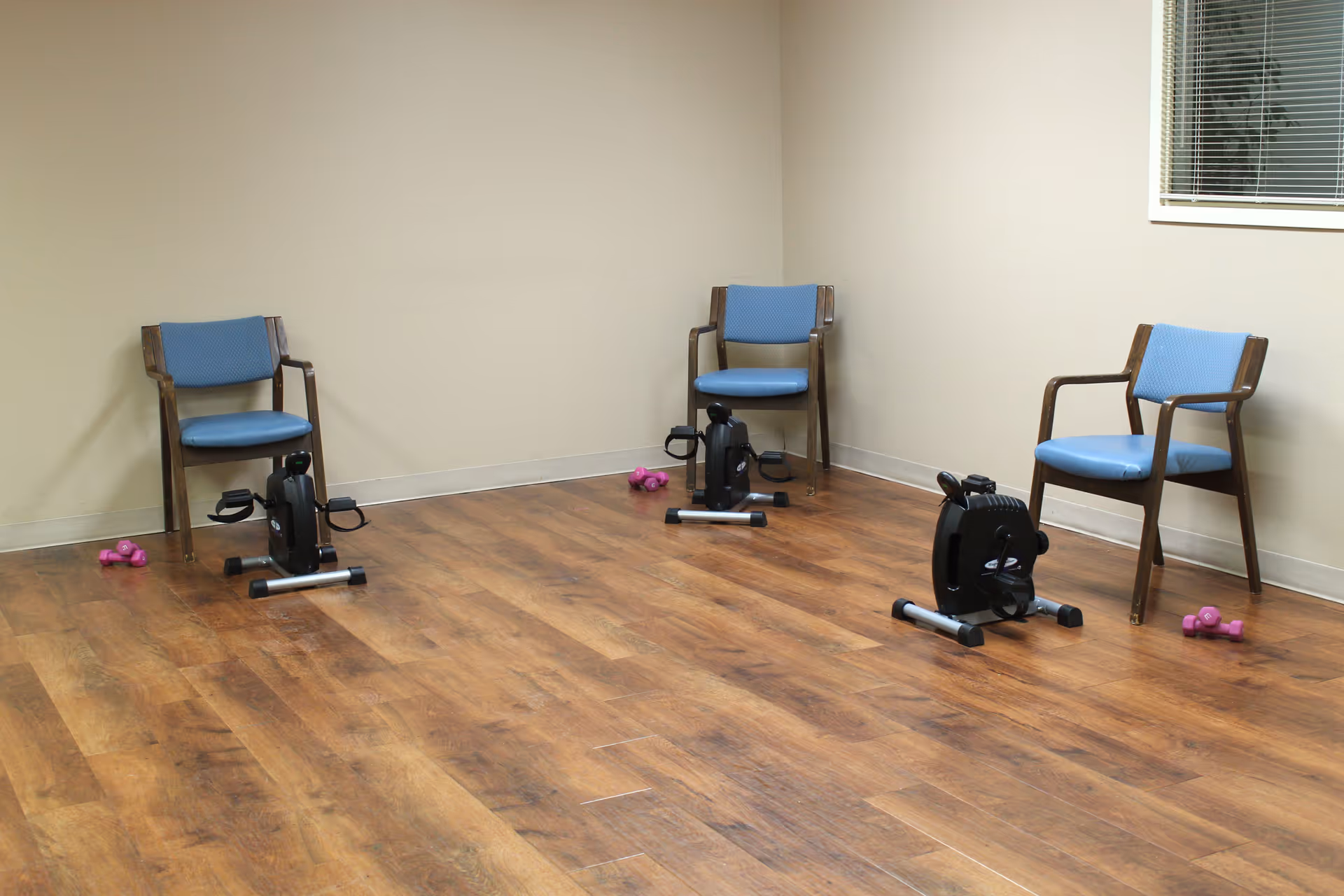 A small exercise room with three wooden chairs with blue cushions, each paired with a mini exercise bike and a set of pink dumbbells on a wooden floor. The walls are beige, and there is a window with blinds on the right side.