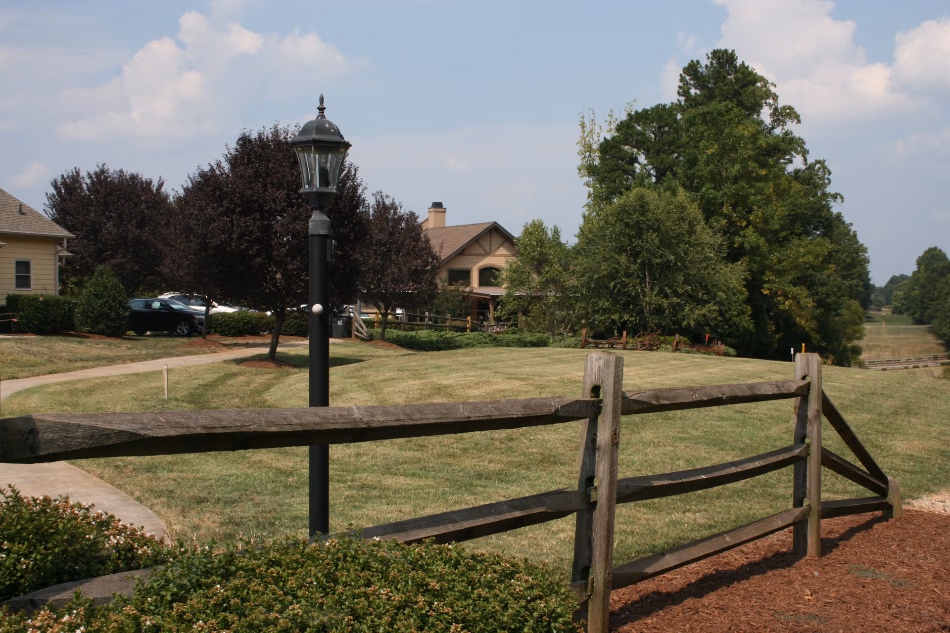 Wooden split-rail fence and a lamp post in front of a grassy lawn with trees and a residential building in the background.
