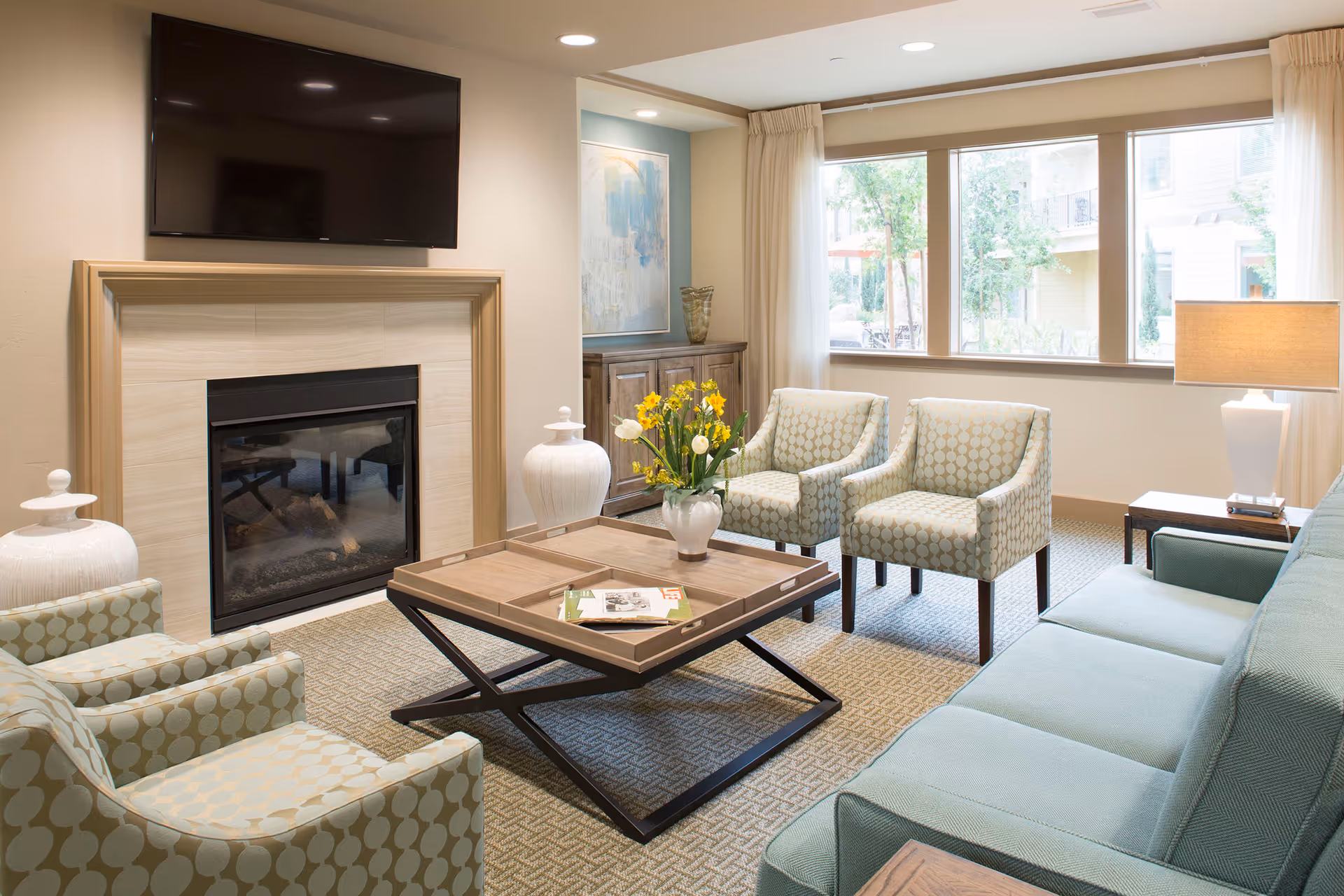 A bright and cozy living room with a large window letting in natural light. The room features a wall-mounted flat-screen TV above a modern fireplace, a wooden coffee table with a vase of yellow and white flowers, two patterned armchairs, a light blue sofa, a side table with a lamp, and a wooden cabinet with decorative items. The carpet has a subtle geometric pattern.
