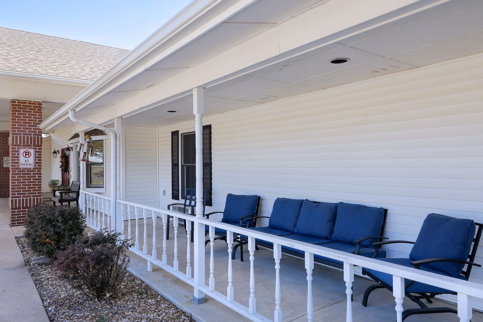 Outdoor covered porch area at Homestead Assisted Living of Russell with white railing, blue cushioned chairs and a bench, and a brick pillar with a no parking sign.