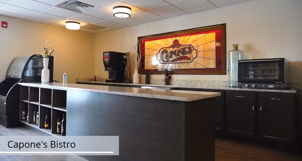 Interior view of a bistro-style kitchen area with a large island countertop, dark cabinetry, a coffee machine, a small oven, and a refrigerated display case. A stained glass window with the text 'Capone's' is mounted on the wall behind the counter. The space has modern ceiling lights and wooden flooring.