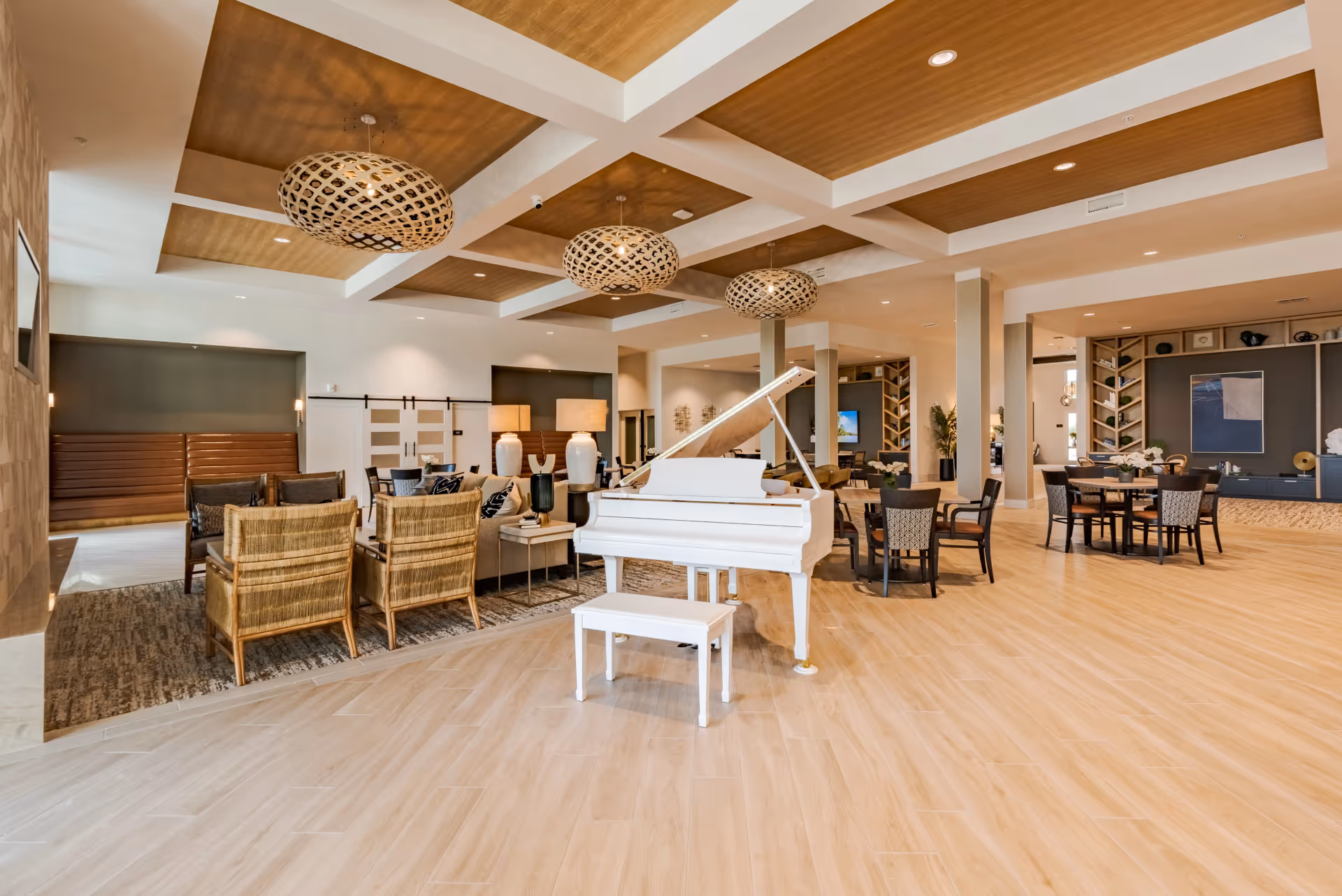Spacious and modern common area with a white grand piano in the center, surrounded by various seating arrangements including wicker chairs, sofas, and dining tables. The room features a coffered ceiling with wooden panels and large decorative pendant lights, light wood flooring, and built-in shelves with decorative items.