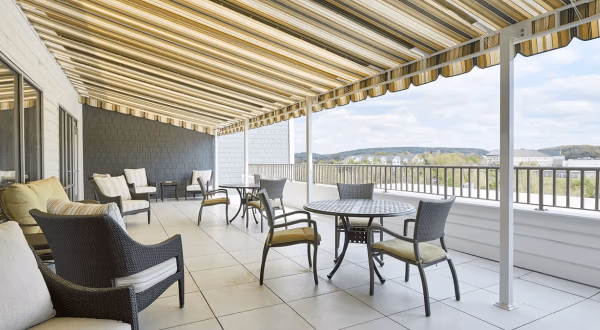 A spacious outdoor covered patio area with several round tables and cushioned chairs arranged for seating. The patio has a striped yellow and white retractable awning overhead and overlooks a scenic view of distant hills and buildings under a partly cloudy sky.