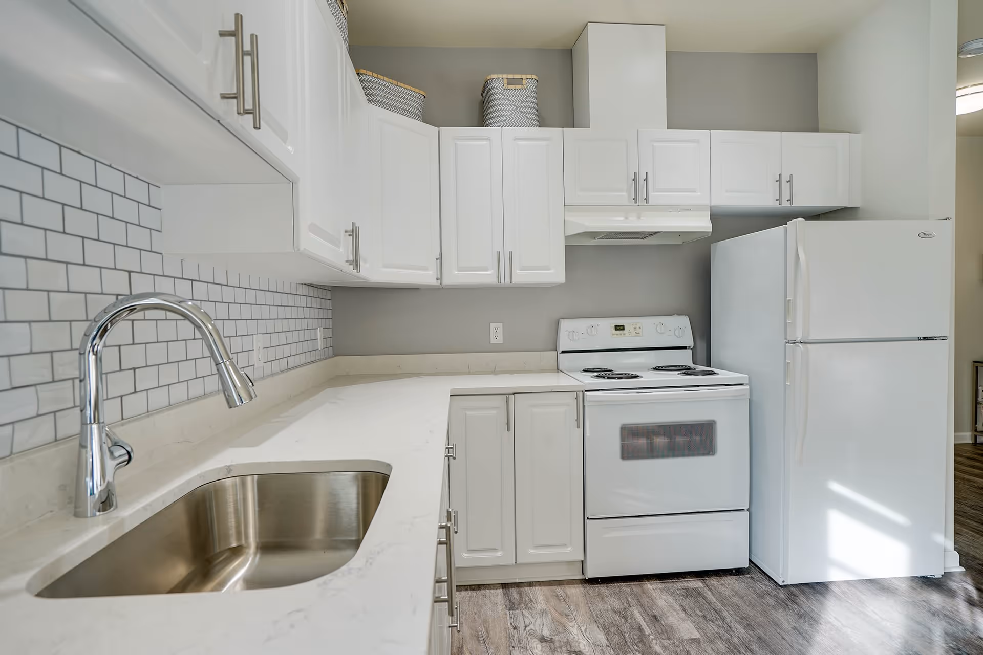 A modern kitchen with white cabinets, a white electric stove, a white refrigerator, a stainless steel sink with a chrome faucet, and a white countertop. The backsplash features white subway tiles with gray grout, and there are two woven baskets on top of the cabinets. The floor has a wood-like finish.