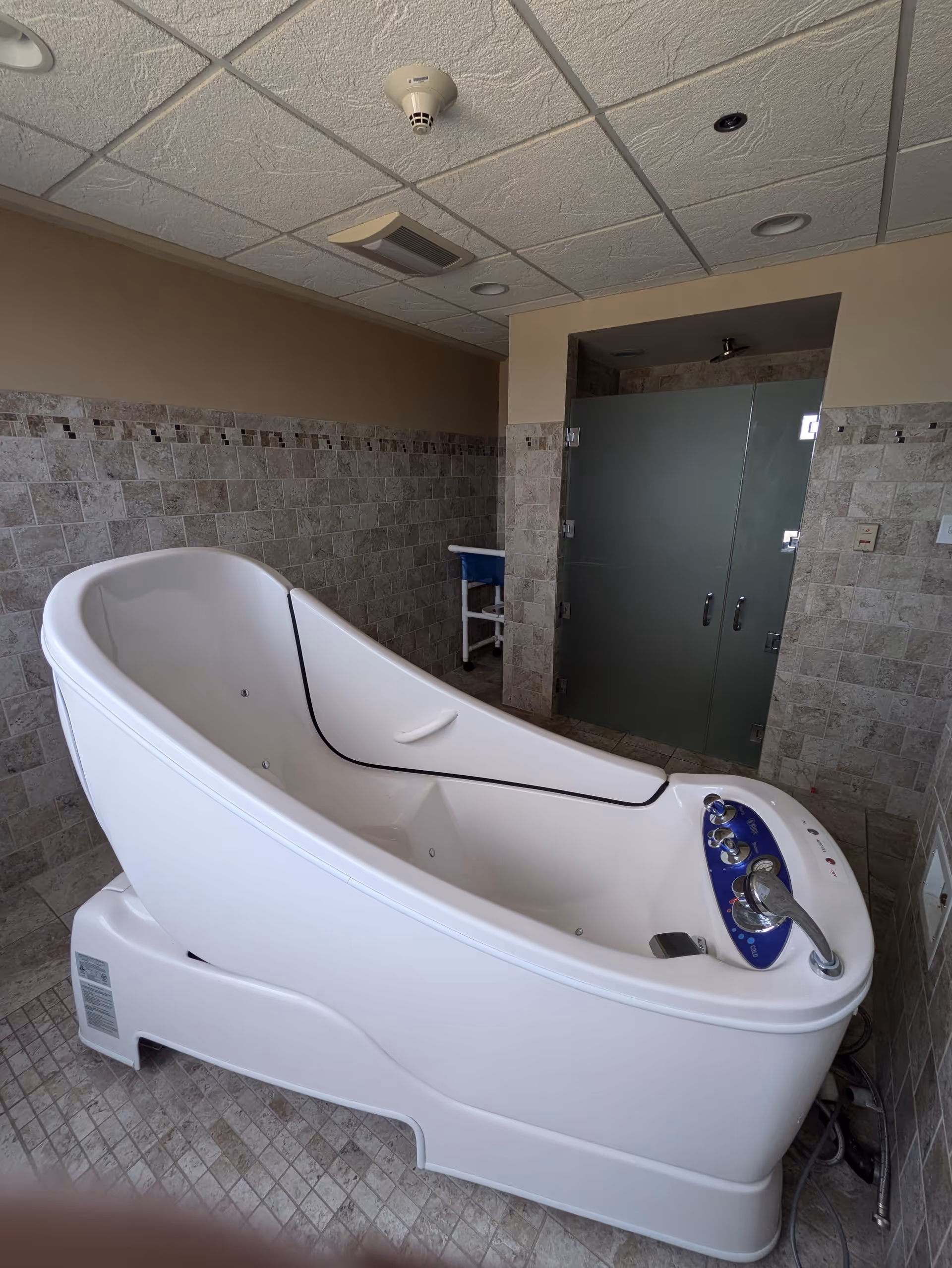A modern white therapeutic bathtub with control knobs and a faucet, situated in a tiled bathroom with a frosted glass shower door in the background.