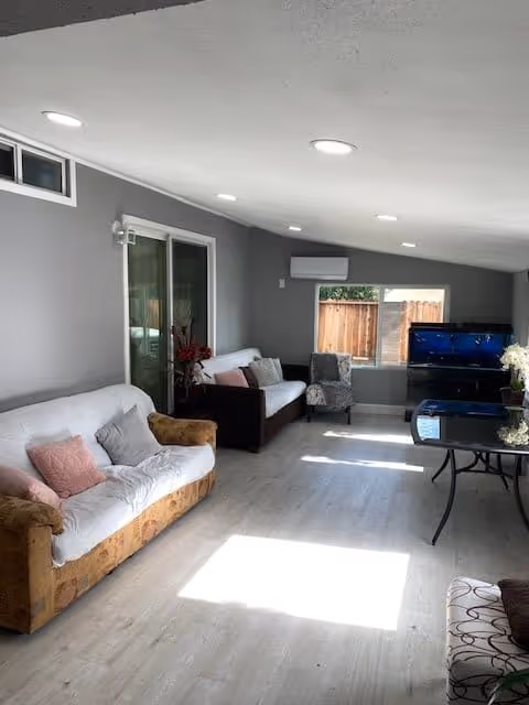 A bright living room with light wood flooring, two sofas with cushions, a patterned armchair, a TV on a stand, a glass table, and a window with a wooden fence visible outside. The walls are painted gray and there are ceiling lights and an air conditioning unit.