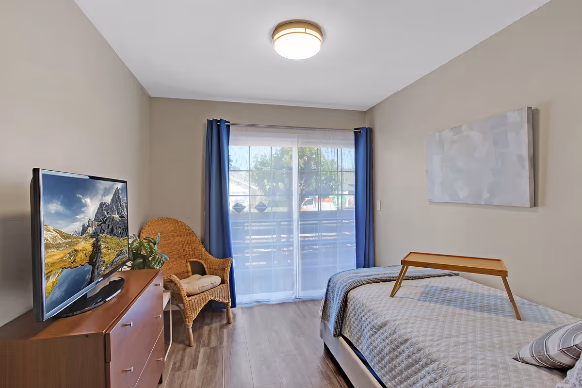 A cozy bedroom with a single bed covered in a light quilt and a small wooden tray on top. There is a wicker chair with a cushion next to a dresser with a flat-screen TV displaying a mountain landscape. The room has a large window with sheer white curtains and blue drapes, allowing natural light to enter. The walls are painted beige, and there is a simple abstract painting hanging above the bed.