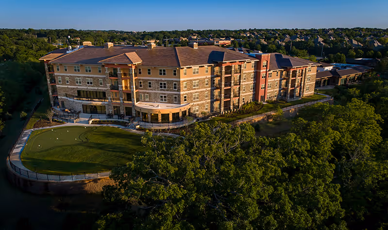 Aerial view of a large multi-story senior living facility building surrounded by trees and greenery, with a putting green area in the foreground and a residential neighborhood in the background under a clear blue sky.