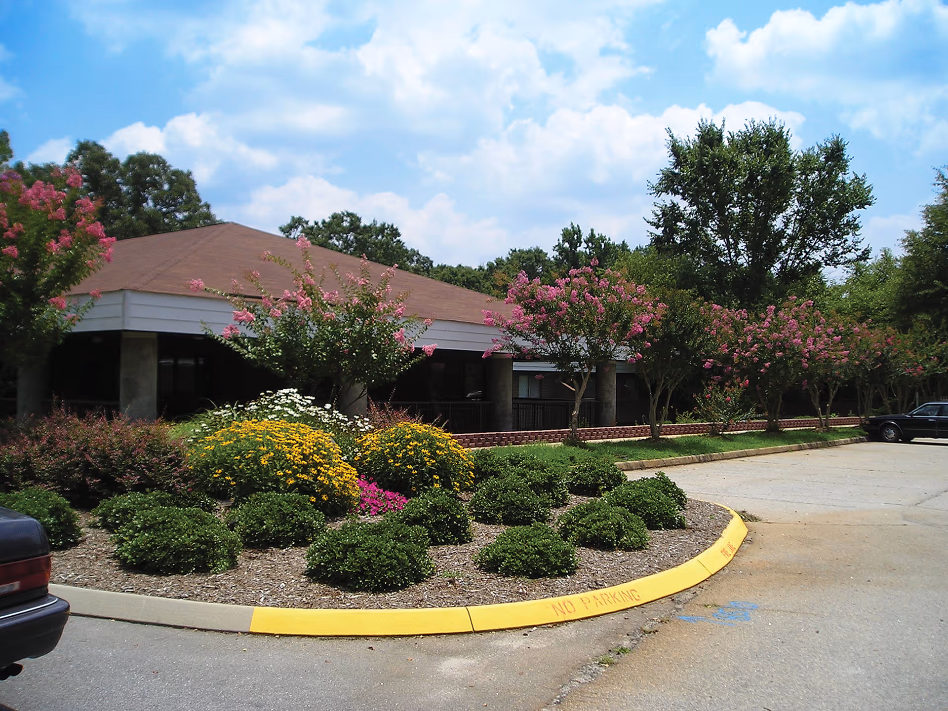 Front exterior of a one-story building with manicured shrubs and pink-flowering trees lining a parking area.