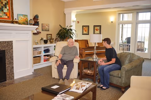 An elderly man and a woman are sitting and talking in a cozy living room area. The man is seated in a beige recliner chair, and the woman is sitting on a green upholstered loveseat. The room features a fireplace with a mantel decorated with various items, a piano against the wall, framed artwork, a table lamp, and a coffee table with magazines and a binder on it. Large windows and a glass door let in natural light.