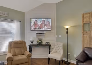 A cozy living room with a beige recliner chair, a white tufted chair, and a dark brown leather chair. A flat-screen TV is mounted on a white shiplap accent wall above a black console table decorated with a flower arrangement and decorative items. A tall floor lamp is lit beside the white chair, and a window with white blinds and curtains is on the left side.