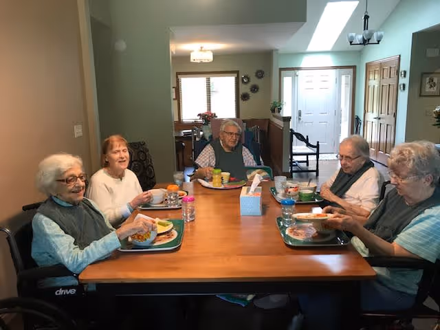 Five elderly individuals sitting around a wooden dining table in a well-lit room, each with a meal tray in front of them. The room has light-colored walls, a window with blinds, and a doorway leading to another area of the facility.