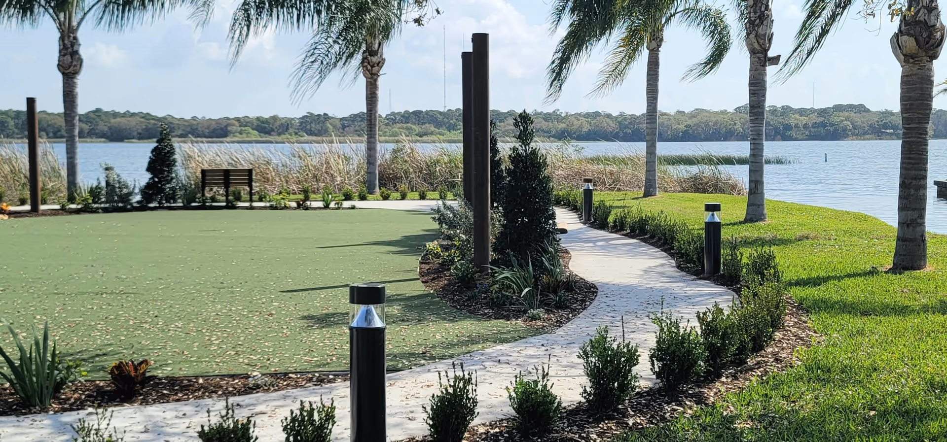 A landscaped outdoor area with a curved concrete pathway lined with small bushes and modern black and silver lamp posts. Palm trees are spaced along the path, and there is a green lawn area with scattered leaves. In the background, there is a large body of water with tall grasses along the shore and a tree line on the opposite bank under a partly cloudy sky.