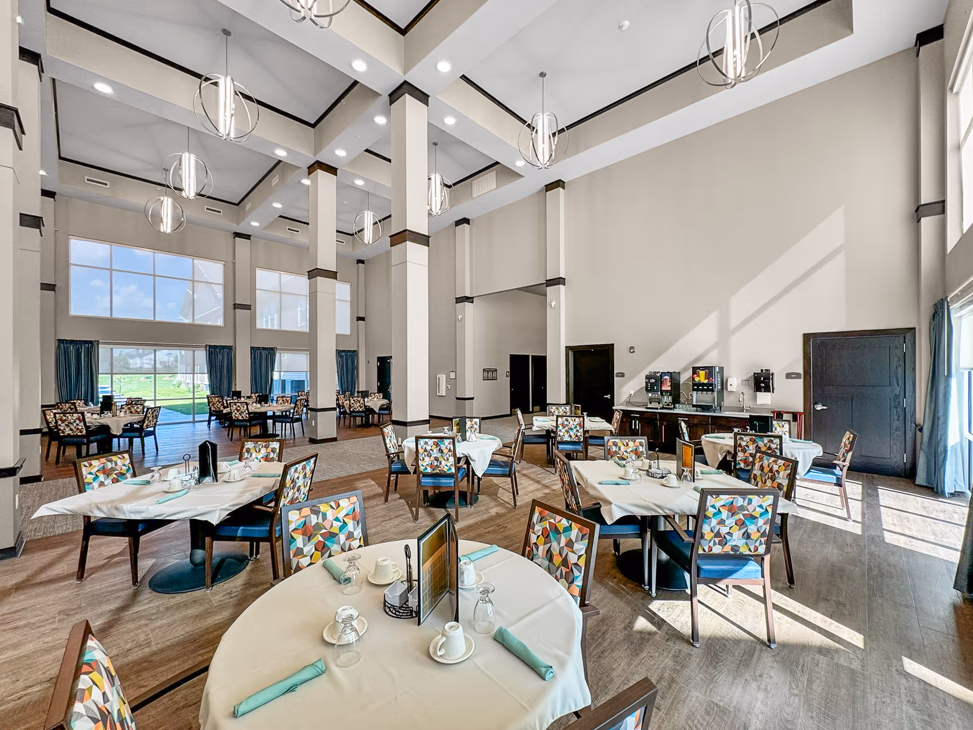 A spacious dining room with high ceilings and large windows allowing natural light to fill the space. Several round and square tables are set with white tablecloths, napkins, cups, and glasses. Chairs with colorful geometric patterned upholstery surround the tables. Modern pendant lights hang from the ceiling, and there is a beverage station against one wall. Blue curtains frame the windows, and the floor is a combination of wood and carpet.