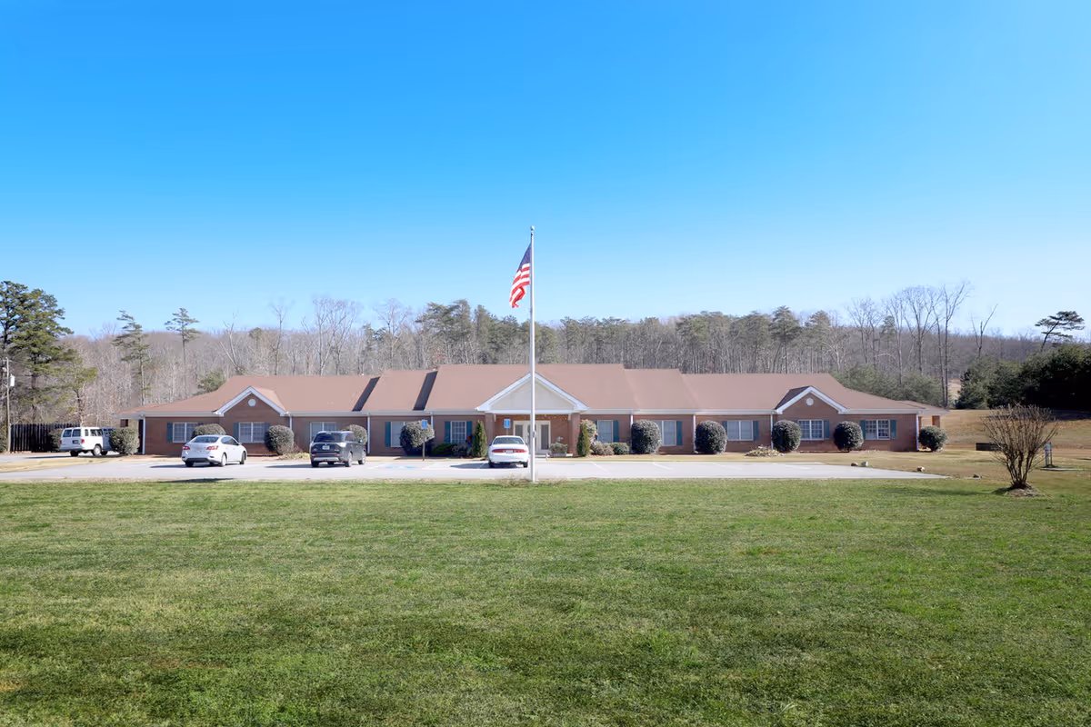 A single-story brick building with a brown roof, several windows with blue shutters, and a central entrance with a white triangular pediment. In front of the building is a flagpole with an American flag, a parking lot with a few cars, and a large grassy area. Trees and a clear blue sky are visible in the background.