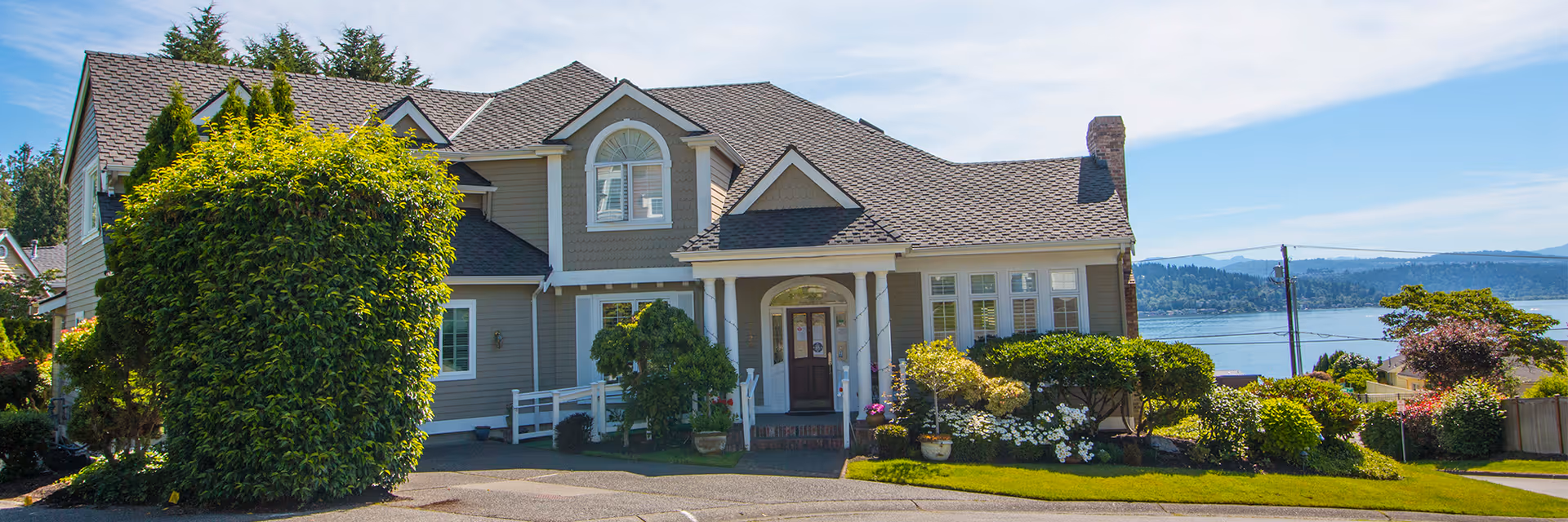 A large two-story house with a gray exterior and white trim, surrounded by well-maintained bushes and greenery. The house has a front porch with columns and a dark front door. In the background, there is a view of a body of water and distant hills under a clear blue sky.
