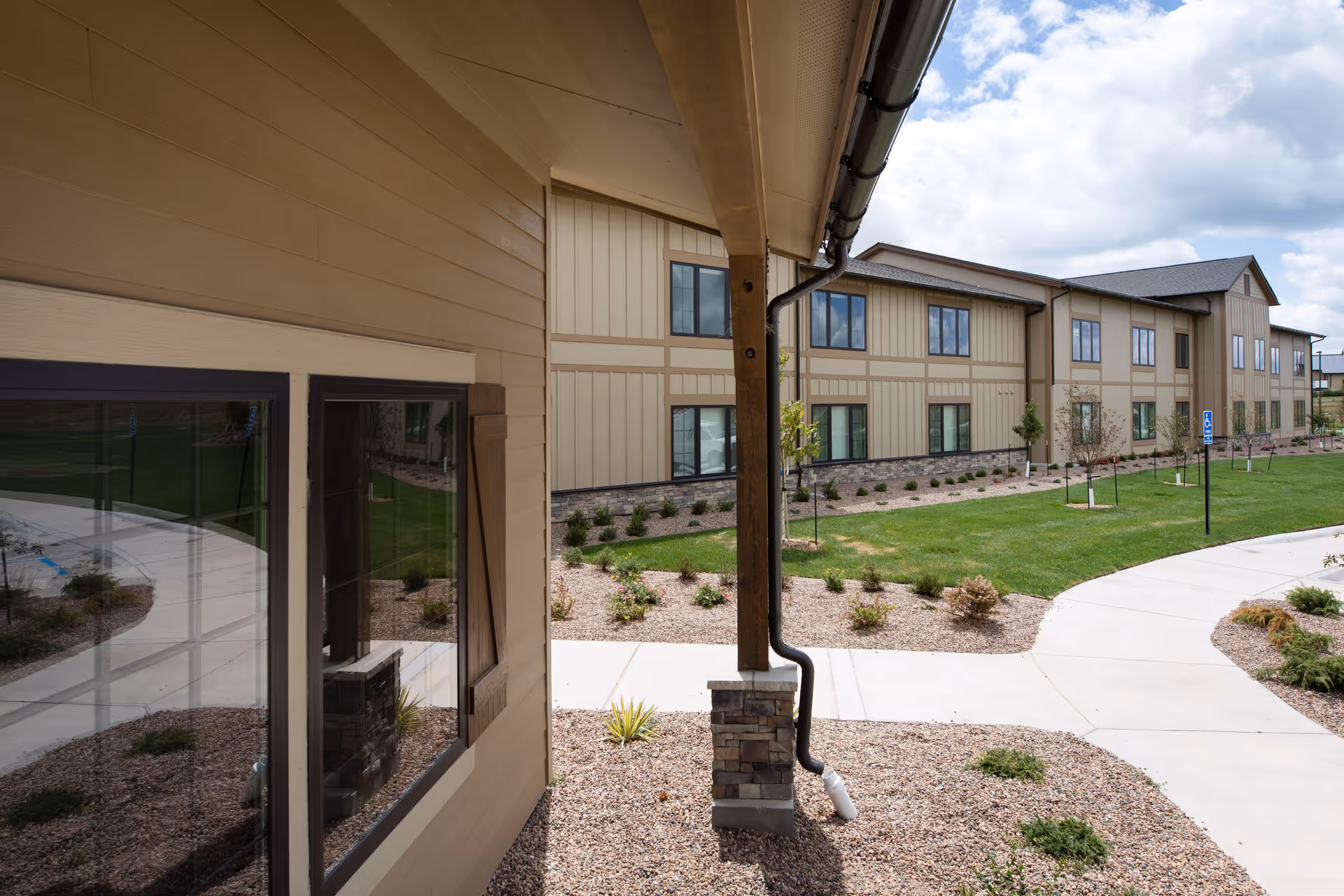 Exterior view of The Ranch House facility showing a two-story building with beige siding and stone accents. The foreground includes a covered walkway with a wooden post and a landscaped area with gravel, small plants, and a curved concrete pathway. The sky is partly cloudy.