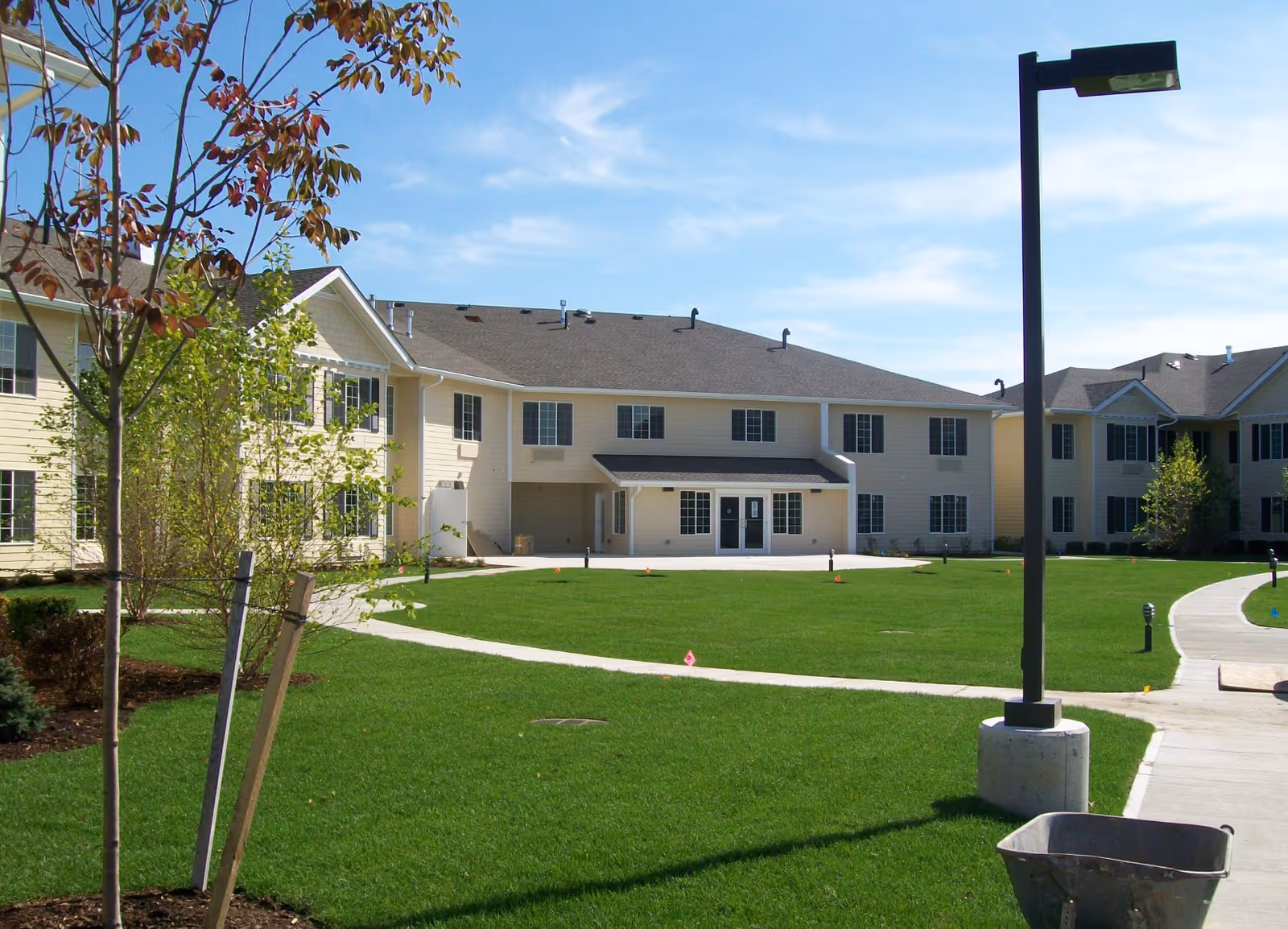 Exterior view of a senior living facility building with beige siding and multiple windows, surrounded by a well-maintained green lawn with young trees and a paved walkway. A tall black street lamp and a wheelbarrow are visible in the foreground under a clear blue sky.