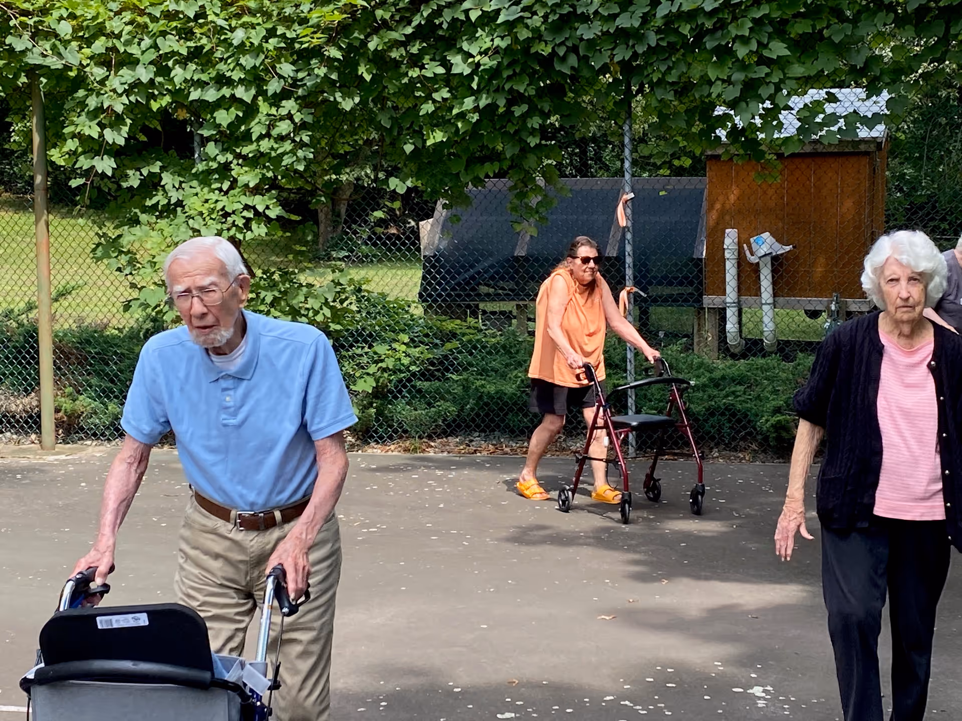 Three elderly individuals outdoors on a paved area near a chain-link fence with greenery and trees in the background. One elderly man in a light blue polo shirt and beige pants is using a walker, a woman in an orange sleeveless top and black shorts is also using a walker, and another woman in a pink shirt and black cardigan is walking nearby.