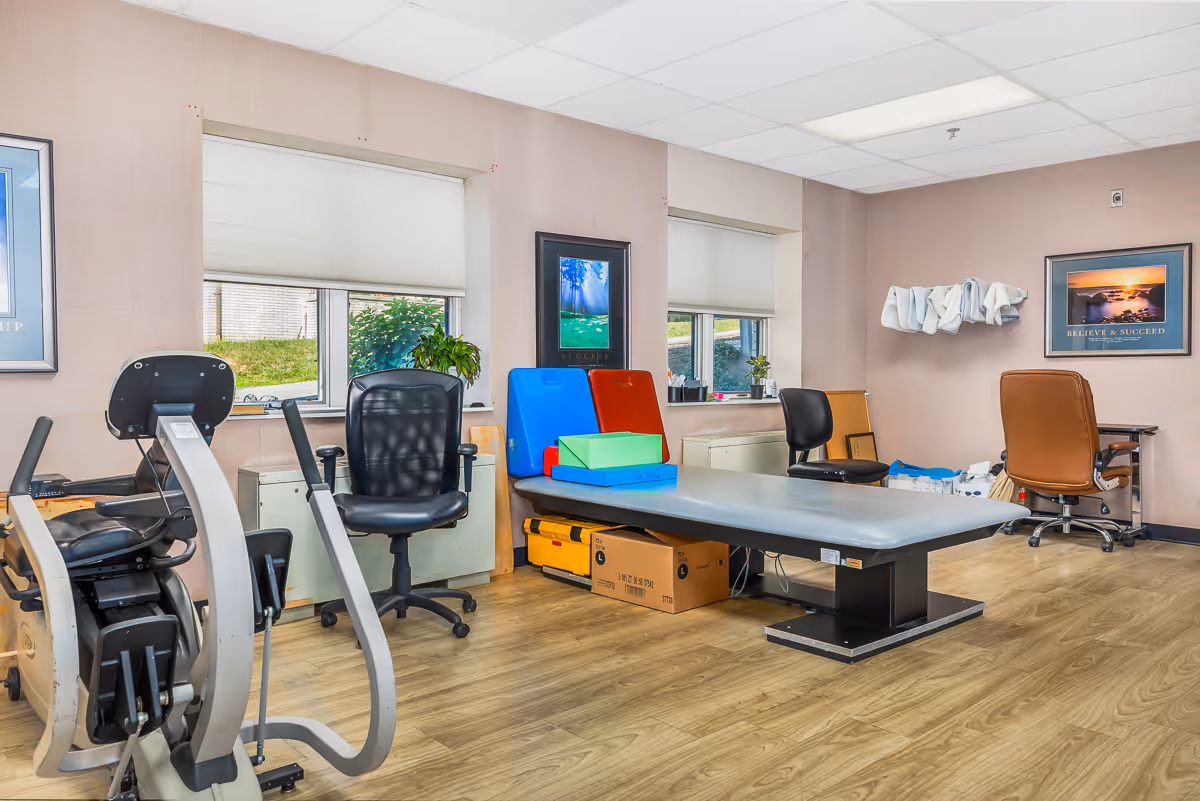 A bright therapy/exercise room with an exercise bike, treatment table, chairs, and colorful therapy mats.