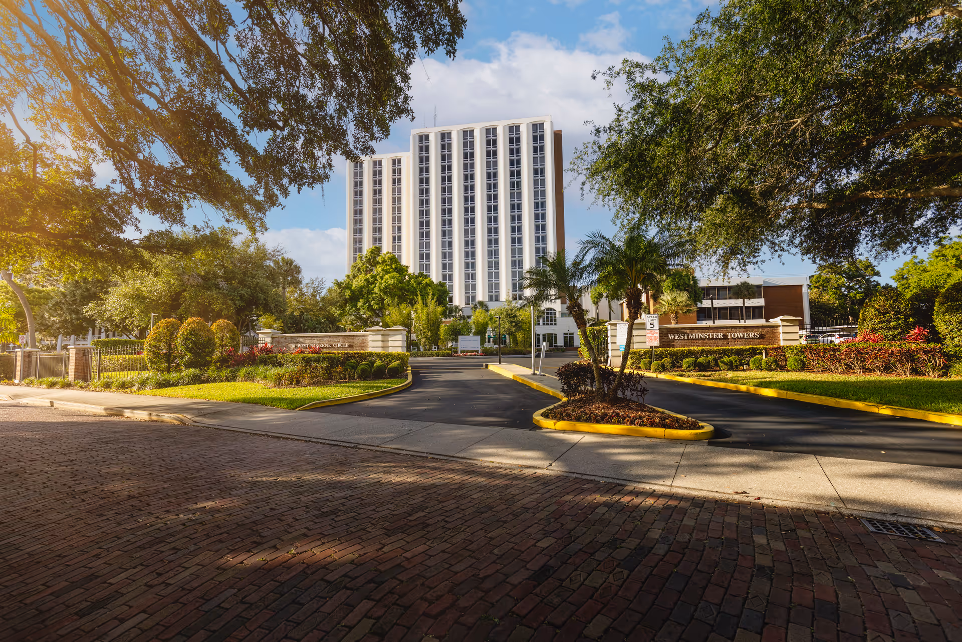 Exterior view of Westminster Towers, a tall multi-story building surrounded by trees and landscaping, with a driveway leading to the entrance and a sign displaying the facility's name.