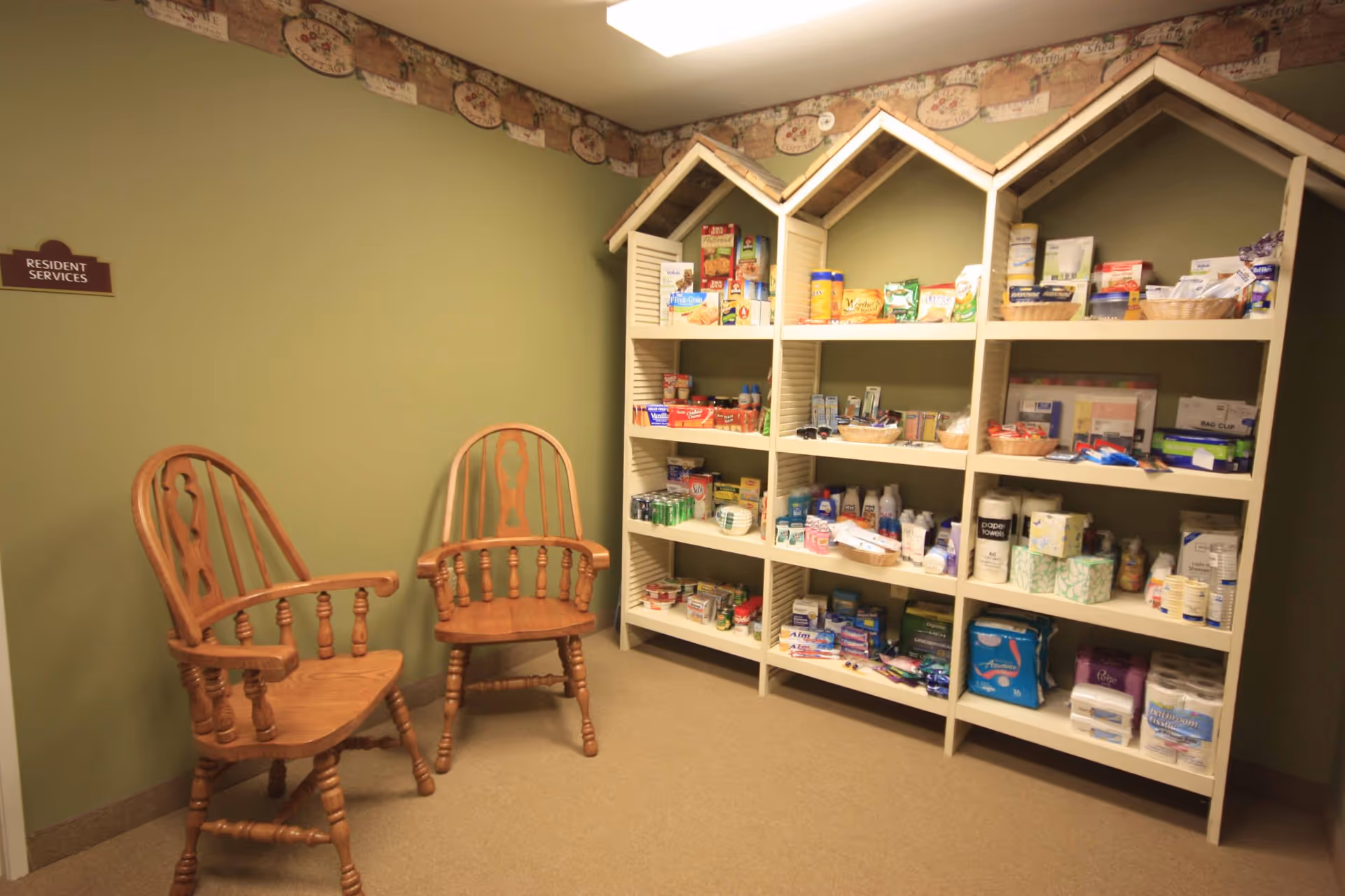 A small room with two wooden chairs and a white shelving unit shaped like houses, stocked with various personal care and household items. The walls are painted green with a decorative border near the ceiling. A sign on the wall reads 'Resident Services'.