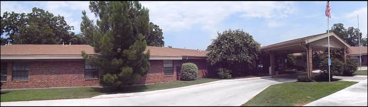 Exterior view of a single-story brick building with a covered entrance and an American flag on a flagpole. The building is surrounded by green grass, trees, and bushes under a partly cloudy sky.