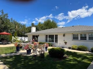 Outdoor patio area of a single-story white building with a gray roof under a blue sky with scattered clouds. The patio features a table with several potted plants and flowers, surrounded by chairs. There is a red umbrella on the left side and green bushes and trees in the background.