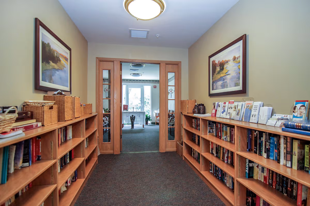 A hallway in Bell Tower Place Senior Apartments with wooden bookshelves filled with books and wicker baskets on both sides. Two framed landscape paintings hang on the beige walls above the bookshelves. At the end of the hallway, there are double wooden doors with glass panels leading to a well-lit room with large windows and some furniture.