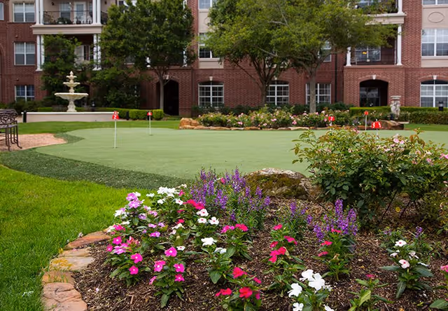 A landscaped outdoor area at The Buckingham featuring a putting green with small red flags, surrounded by green grass, colorful flower beds with pink, white, and purple flowers, and trees. In the background, there is a multi-story brick building with windows and balconies.