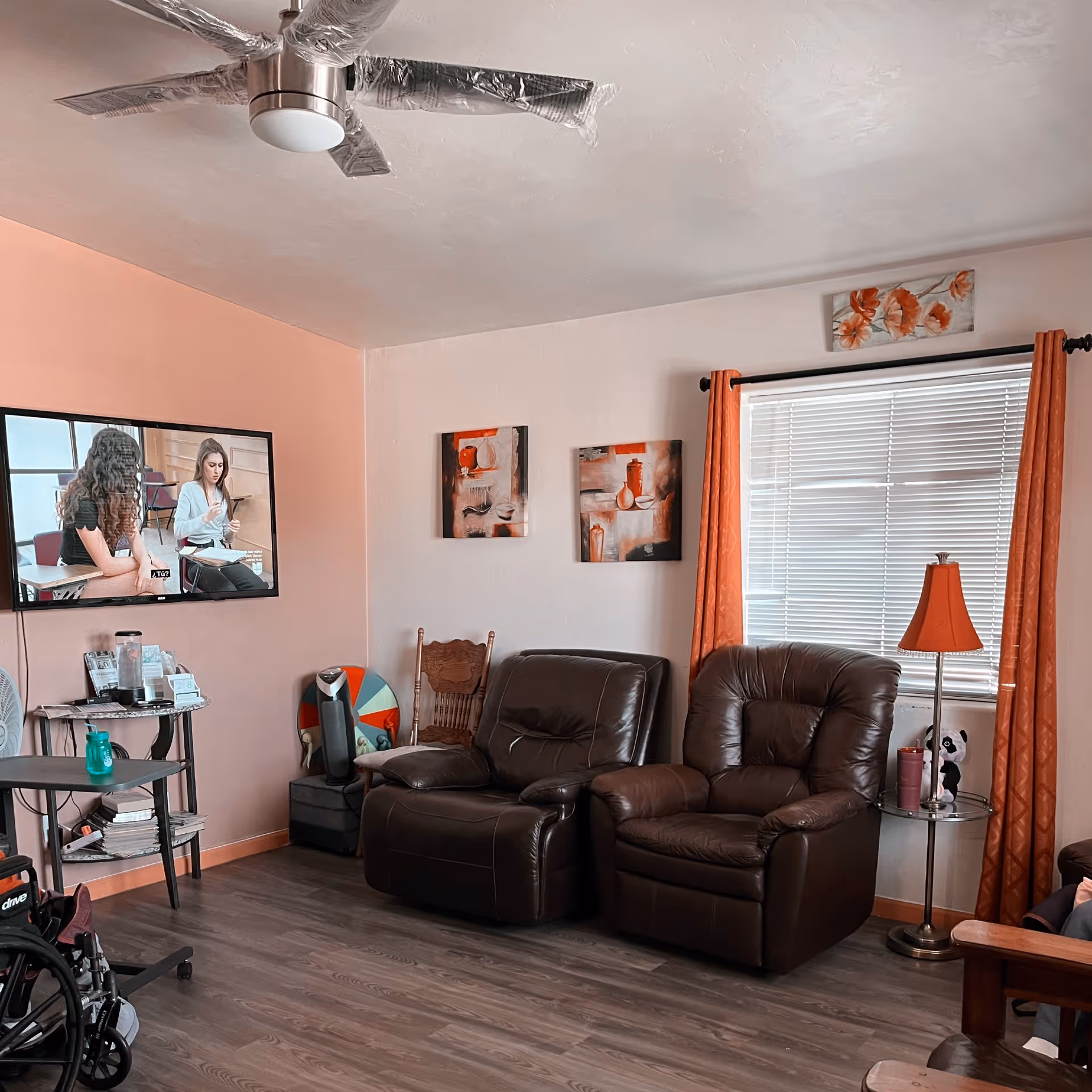 Living room with two brown leather recliners facing a wall-mounted TV, orange curtains, and hardwood floors.