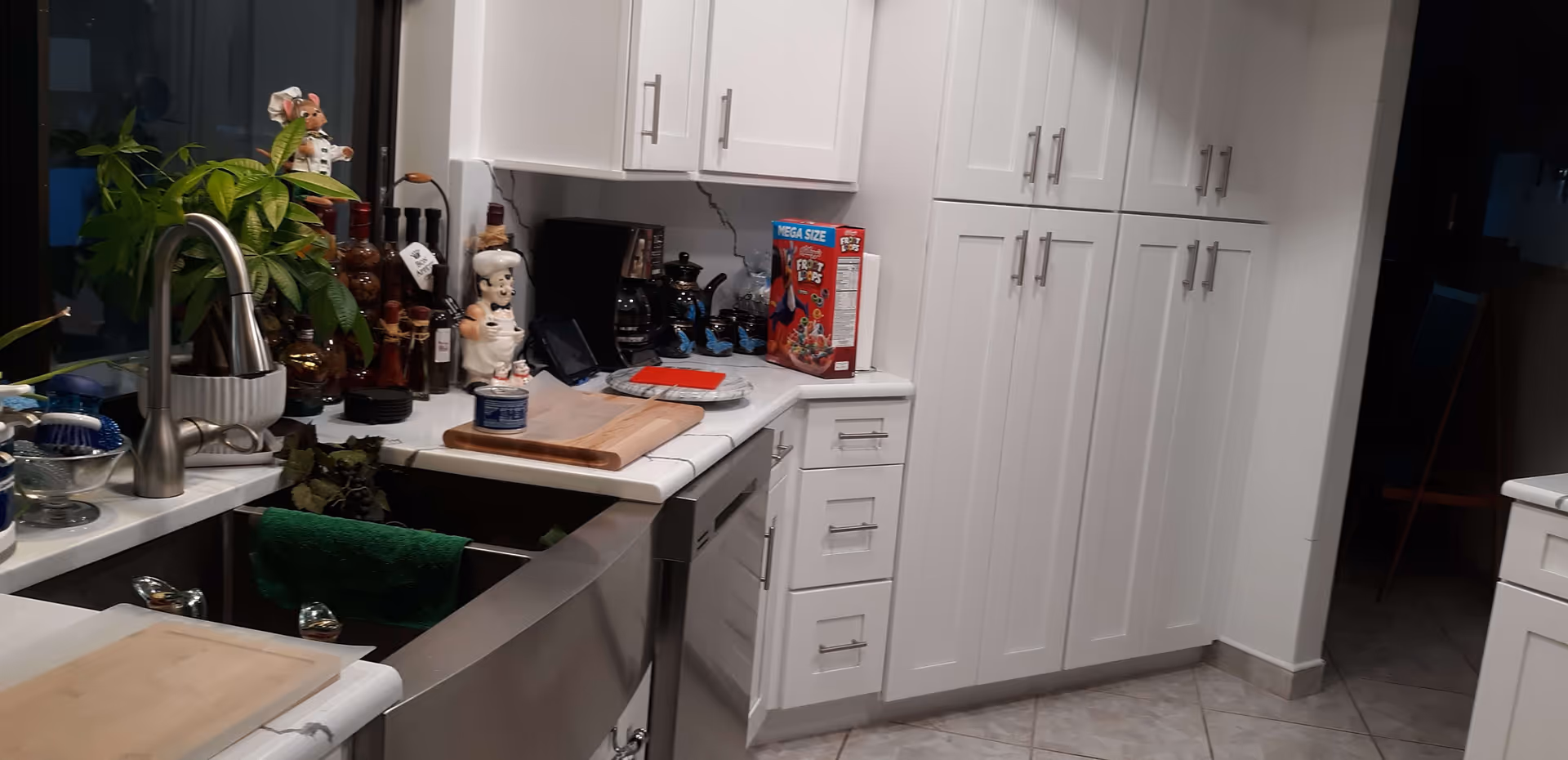 A modern kitchen with white cabinets and drawers, a stainless steel sink with a green towel hanging on it, a variety of kitchen items on the countertop including a box of Froot Loops cereal, a coffee maker, decorative chef figurines, and a potted plant near the window.