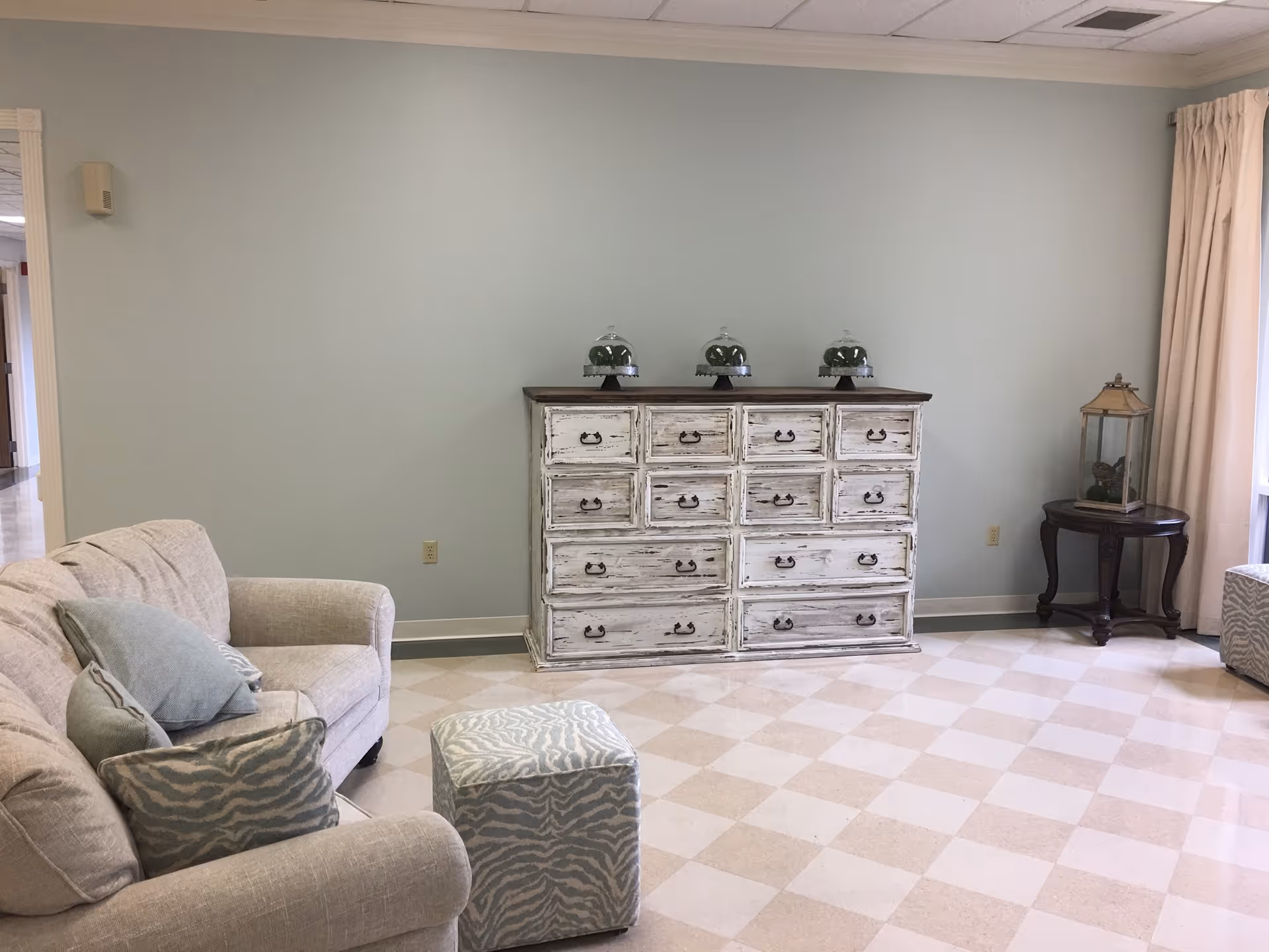 A cozy living room area with a beige couch adorned with patterned pillows, a matching ottoman, a distressed white wooden dresser with multiple drawers, and a small round wooden side table with a decorative lantern. The room has light-colored checkered flooring and pale blue walls with a window covered by beige curtains.