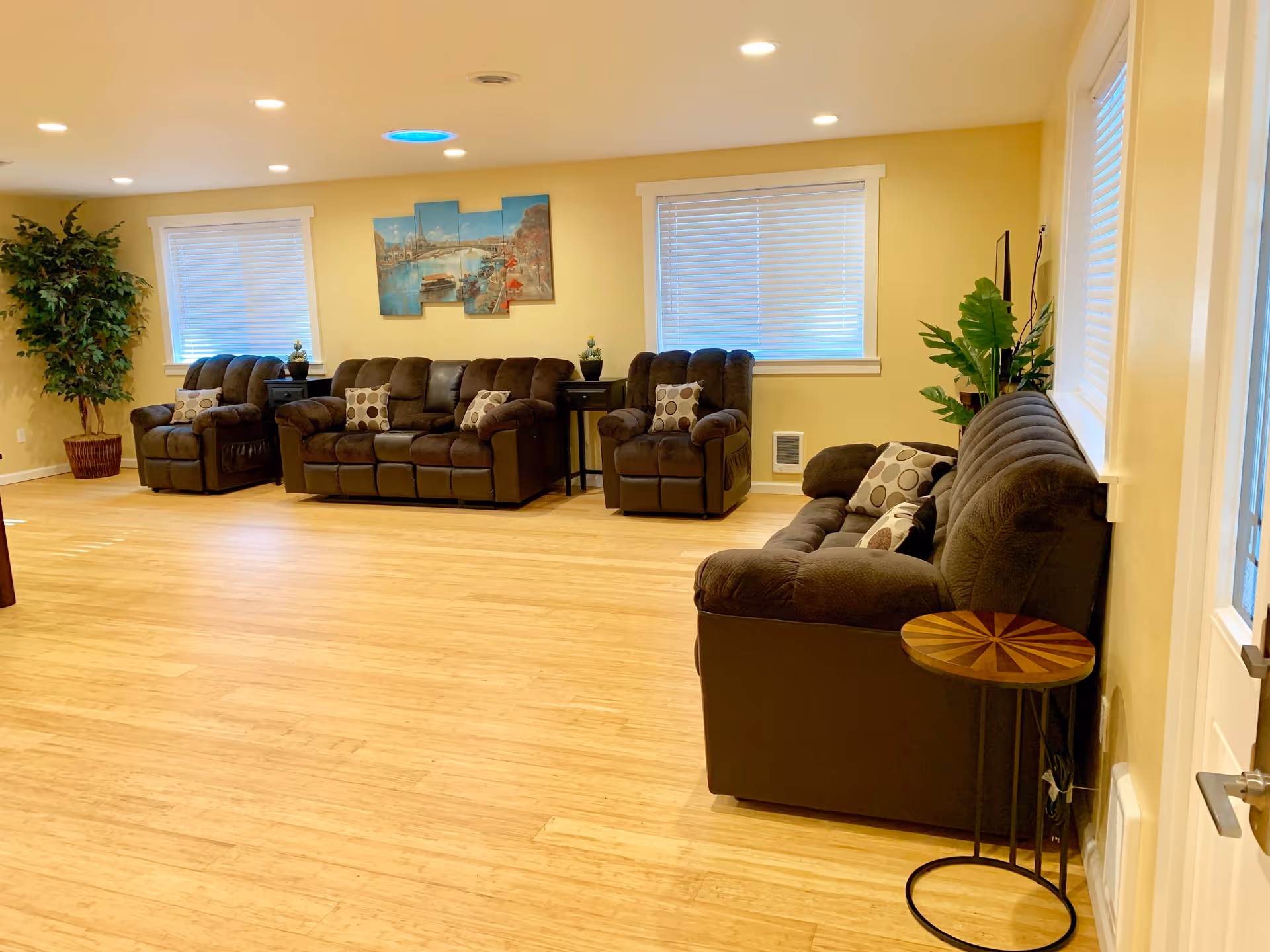 A spacious living room with light yellow walls and wooden flooring. The room features three brown cushioned sofas with patterned pillows, two windows with white blinds, a small round wooden side table, two black side tables with small plants, a large potted plant in the corner, and a multi-panel wall art depicting a river scene.